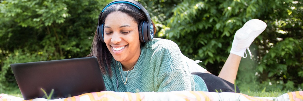 This image depicts a young adult on a blanket in the grass using a laptop with headphones.